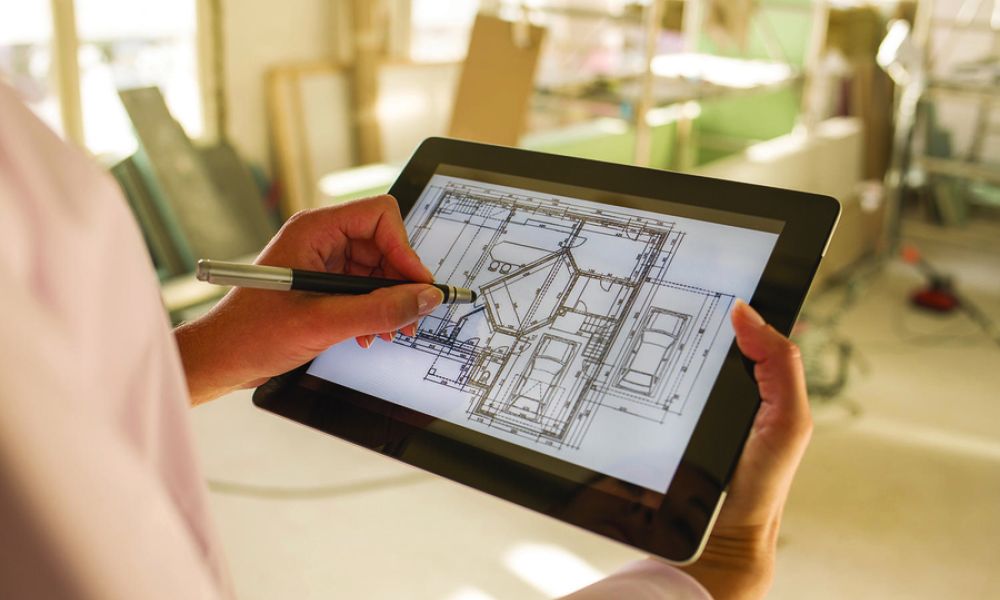 A person using a stylus on a tablet displaying architectural blueprints, set in a construction environment with building materials and scaffolding in the background.