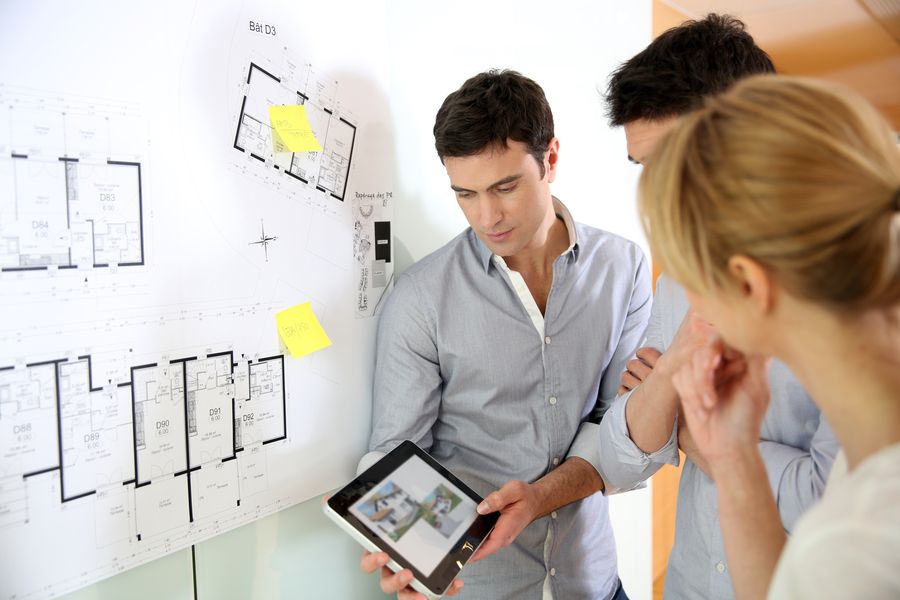 A professional reviews architectural floor plans pinned to a wall with a couple, while he holds a tablet displaying a digital rendering.
