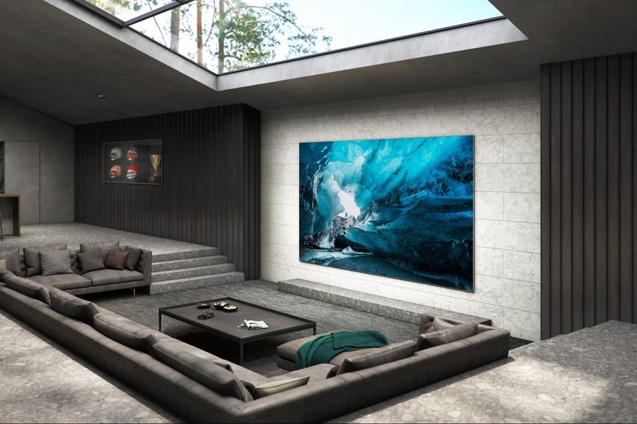 A modern luxury living room with a large video wall display showing an image of an icy cave, surrounded by sleek gray furniture, stone floors, and a skylight ceiling