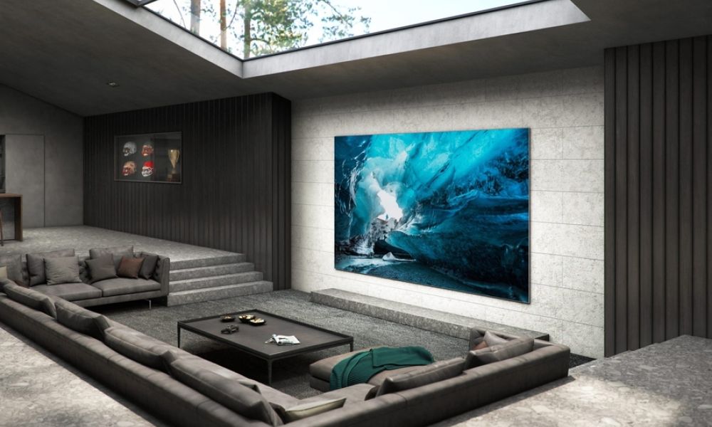 A modern luxury living room with a large video wall display showing an image of an icy cave, surrounded by sleek gray furniture, stone floors, and a skylight ceiling