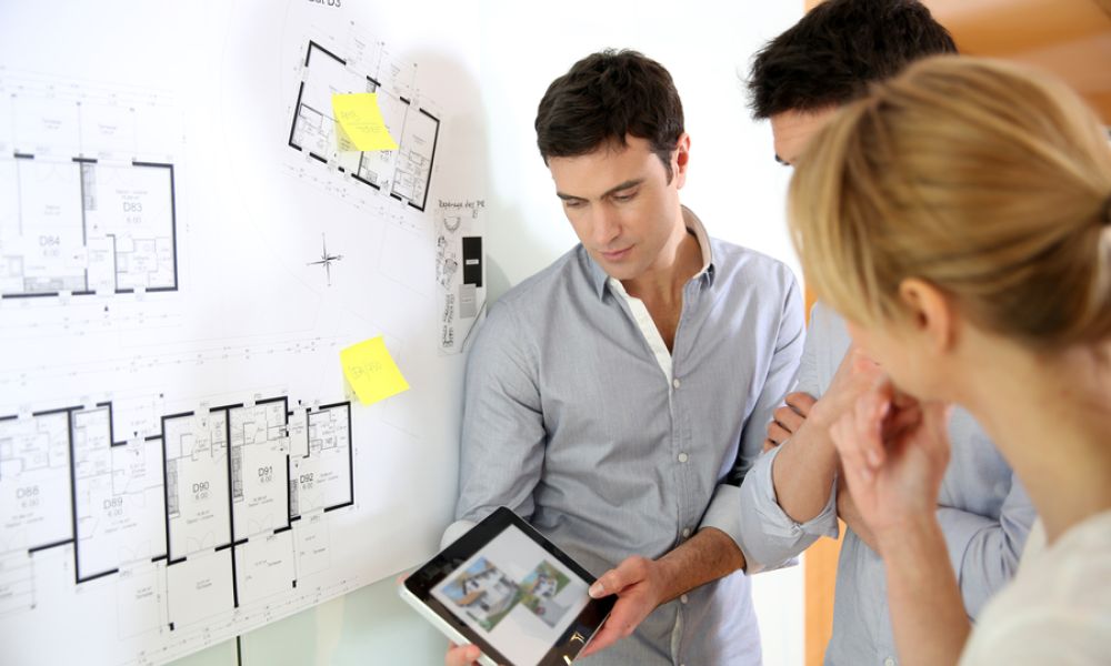 A professional reviews architectural floor plans pinned to a wall with a couple, while he holds a tablet displaying a digital rendering.